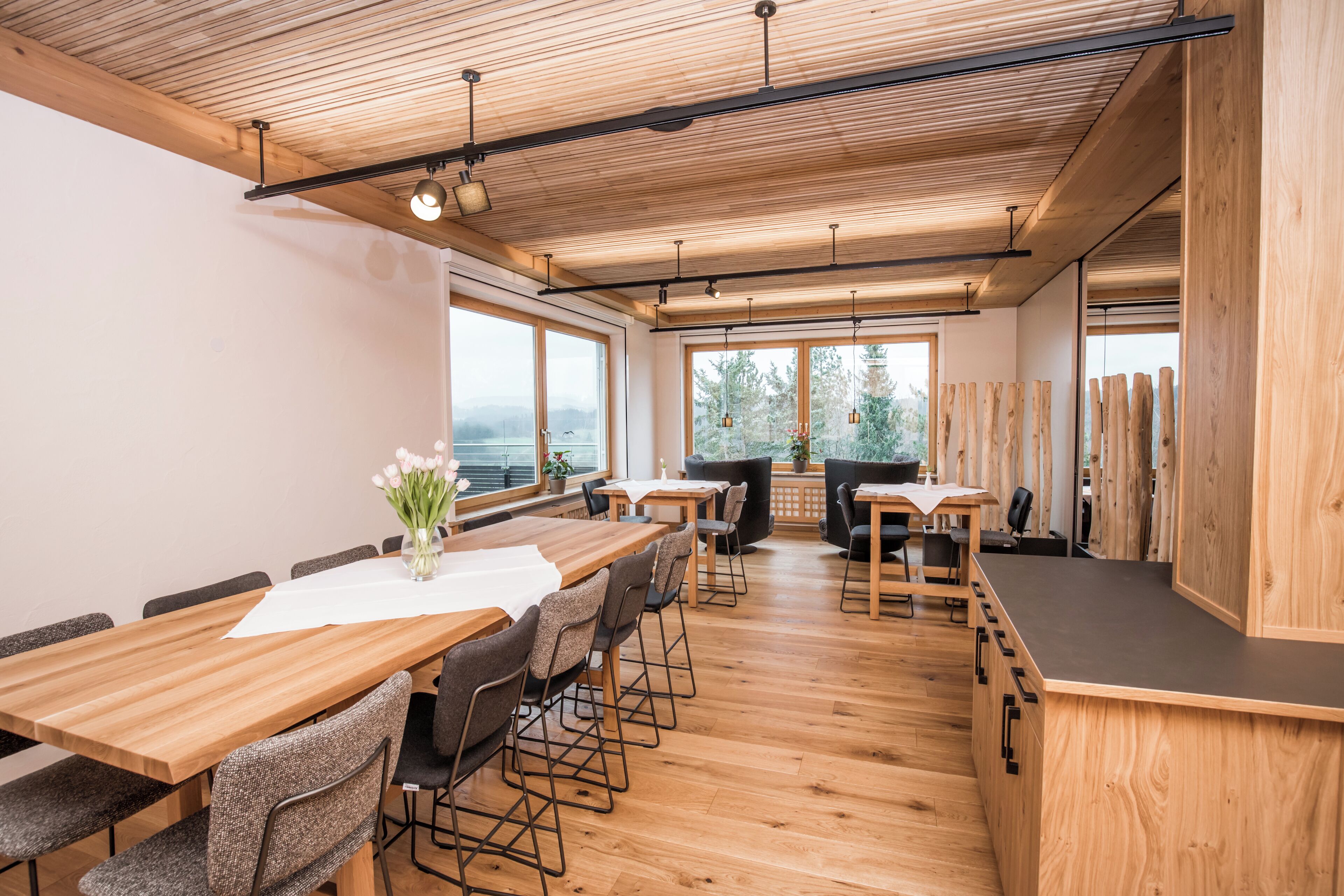 Ein modernes Interieur mit Holztischen und -stühlen, großen Fenstern und Blick ins Freie.