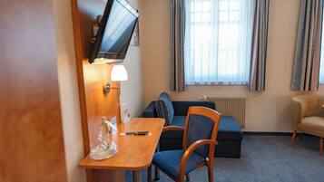 A hotel room with a wooden desk, a television mounted on the wall, a sofa, and a chair.