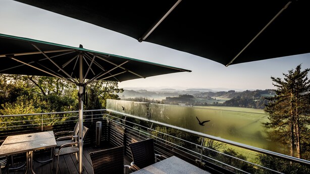 Una terraza exterior con una mesa y sillas, una barandilla de metal y vistas a un lago y montañas distantes.