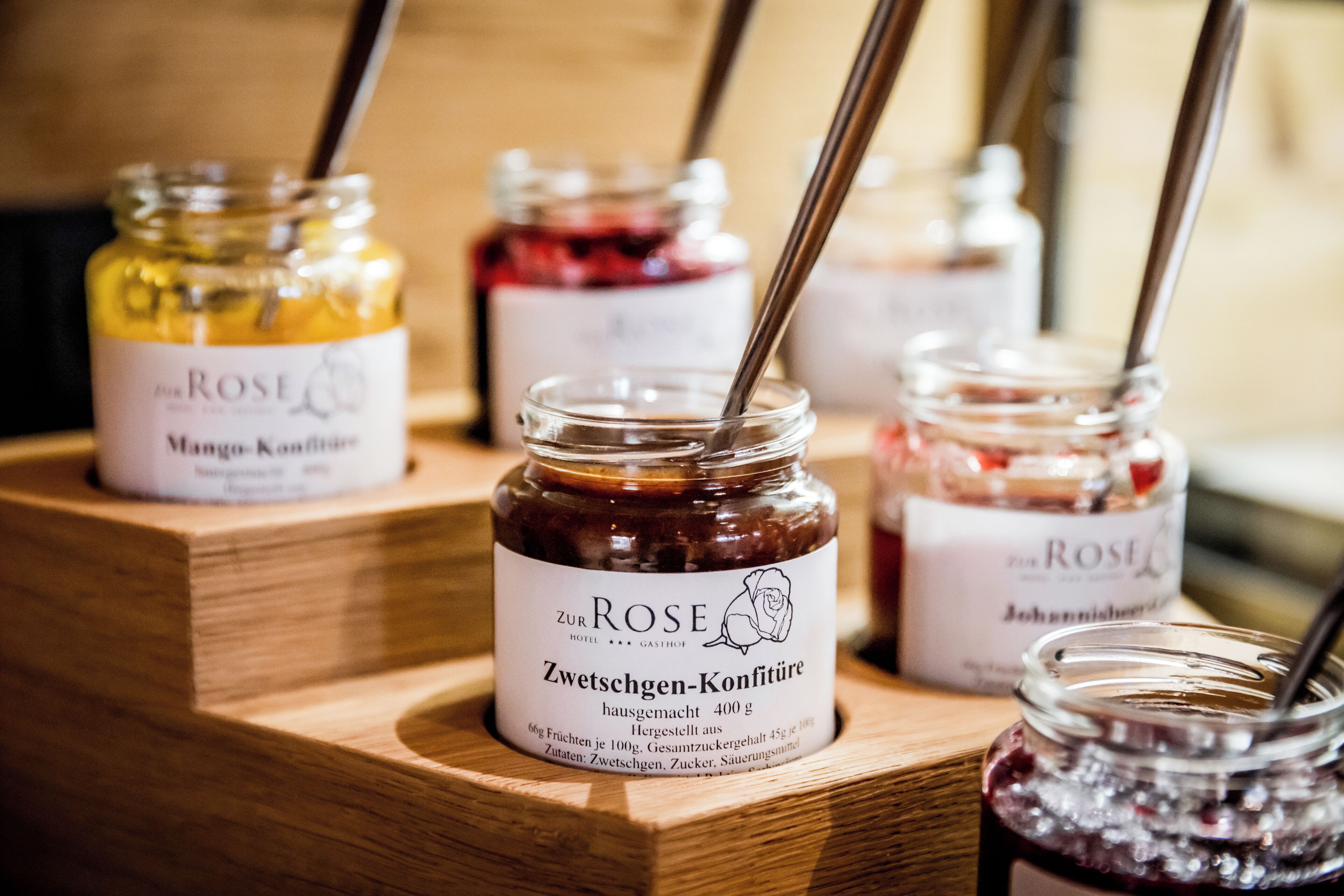 Uma apresentação de madeira com vários potes de geleia, cada um etiquetado com o nome 'Zur Rose' e diferentes sabores como 'Mango-Konfitüre' e 'Johannisbeersirup'.