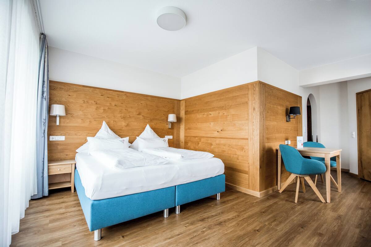 A hotel room with a wooden accent wall, a bed with white bedding, a desk with a chair, and a lamp.