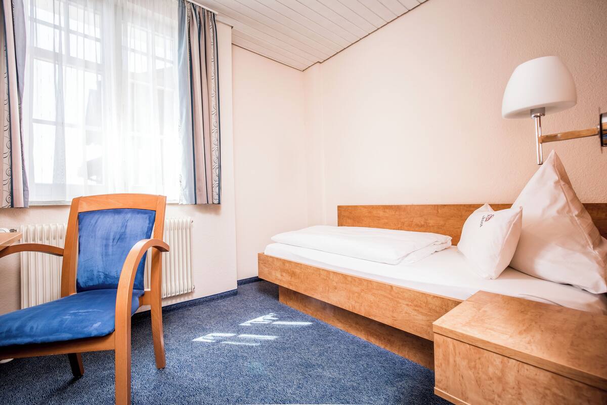 A hotel room with a wooden bed, a blue chair, a white lamp, and a window with curtains.