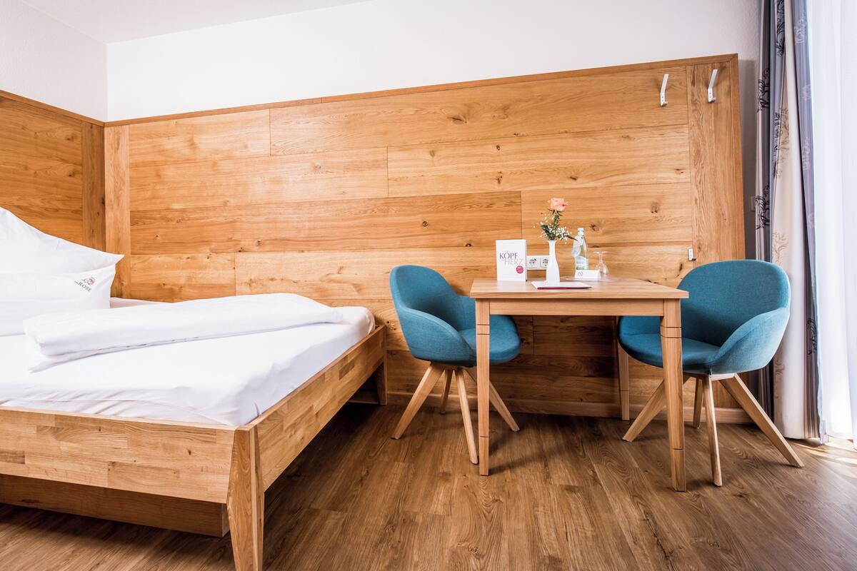A wooden bed with white linens, a small wooden desk with two blue chairs, and a wooden wardrobe.
