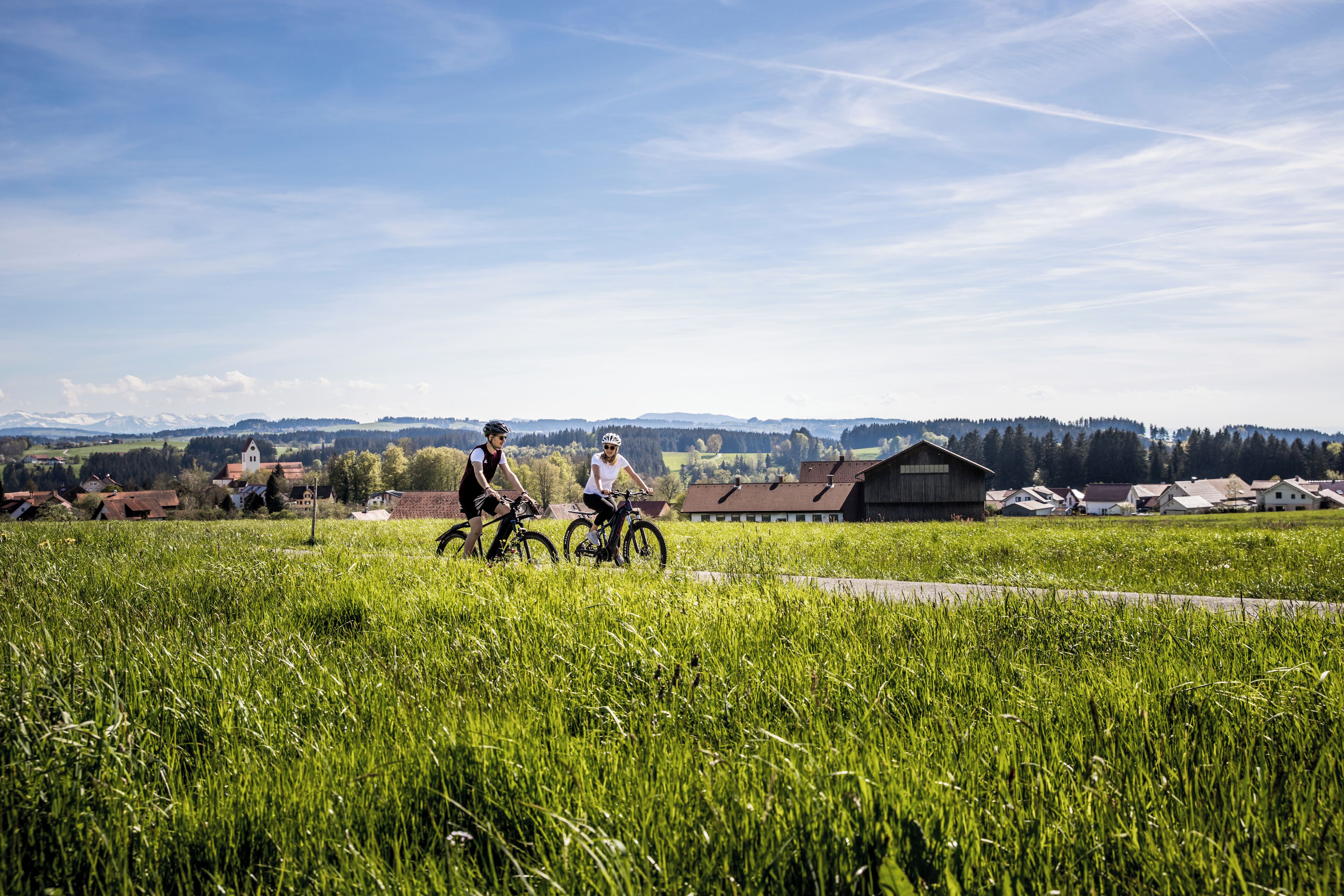 Two cyclists on a rural road with houses and a barn in the background.