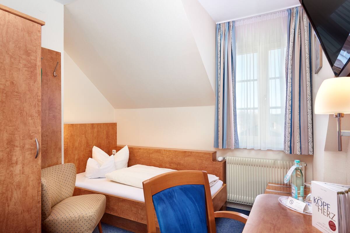 A hotel room with a bed, a chair, a desk, a radiator, and a window with curtains.