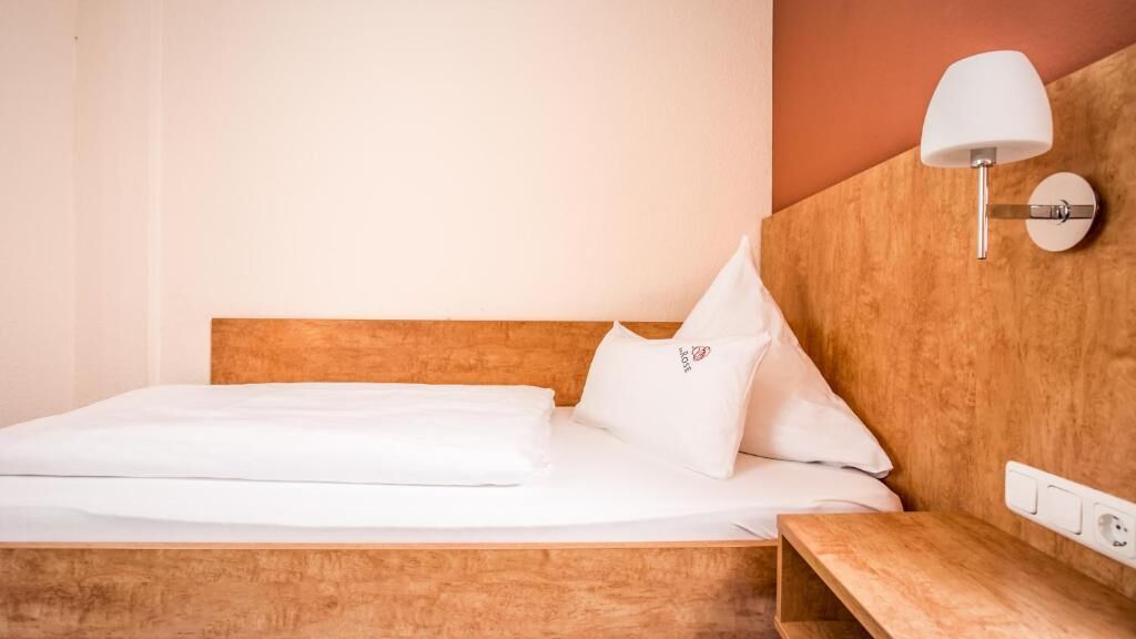 A neatly made bed with white linens, a wooden headboard, a bedside table with a lamp, and a wall socket.