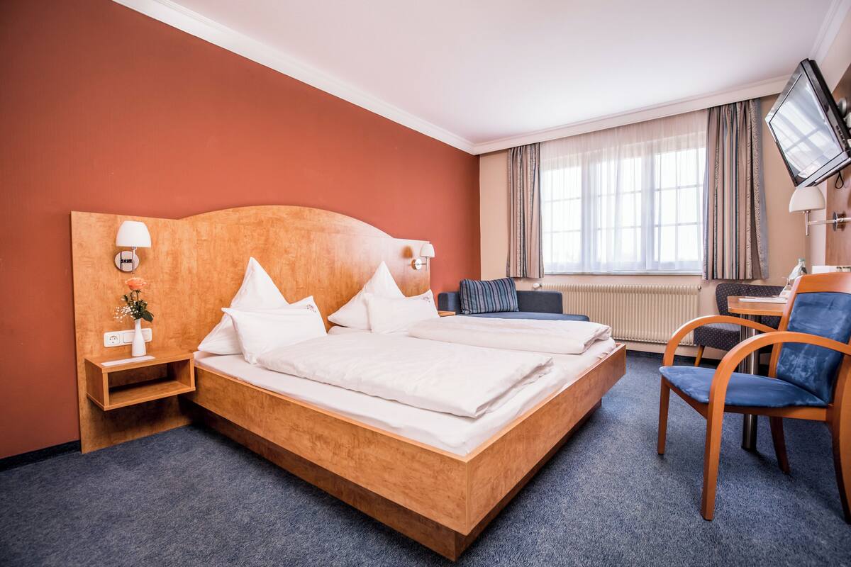 A hotel room with a wooden bed, a desk, a chair, a TV, and a window with curtains.