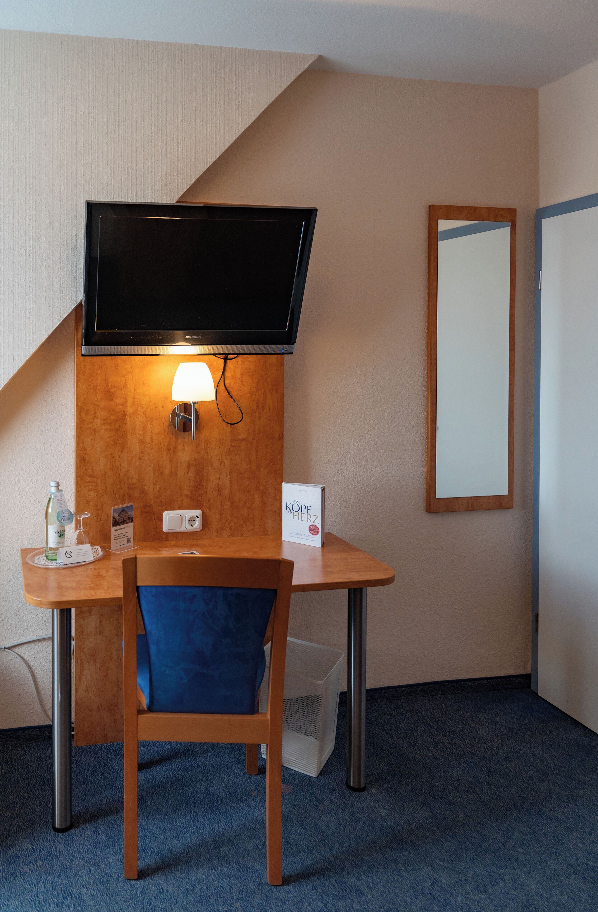 Een hotelkamer met een houten bureau, een blauwe stoel, een flatscreen televisie aan de muur, een wandlamp en een spiegel.