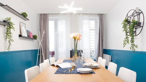 A dining area with a wooden table set for four, surrounded by white chairs. There are shelves with plants and decorative items on the walls, and a large window with curtains. - Elegant, Air-conditioned & Central, close Marais (Paris)