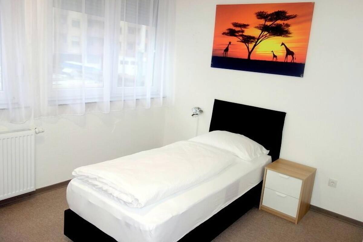 A neatly made bed with a white comforter and pillows, a small bedside table with a drawer, a wall-mounted painting of a sunset with giraffes, and a window with sheer curtains.