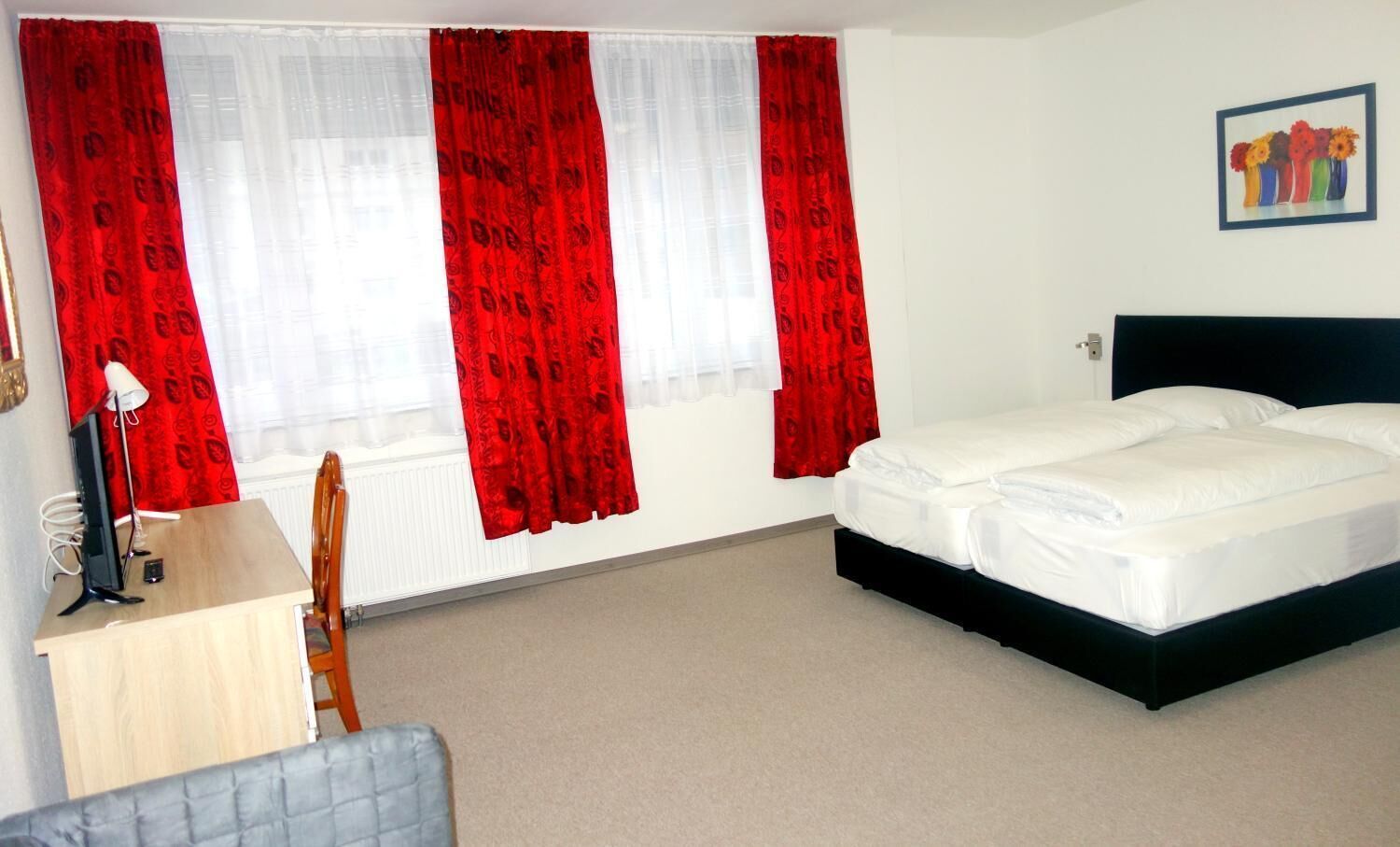 Habitación de hotel con cama, escritorio con lámpara, una silla y ventana con cortinas rojas.