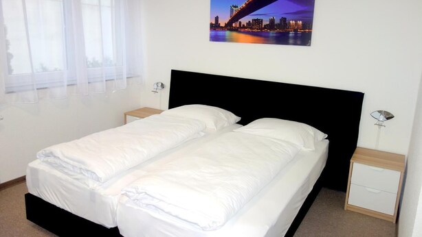 A double bed with white bedding, two bedside tables, a nightstand, a window with sheer curtains, and a cityscape picture on the wall. - Budget Hotel Ludwigshafen (Ludwigshafen)