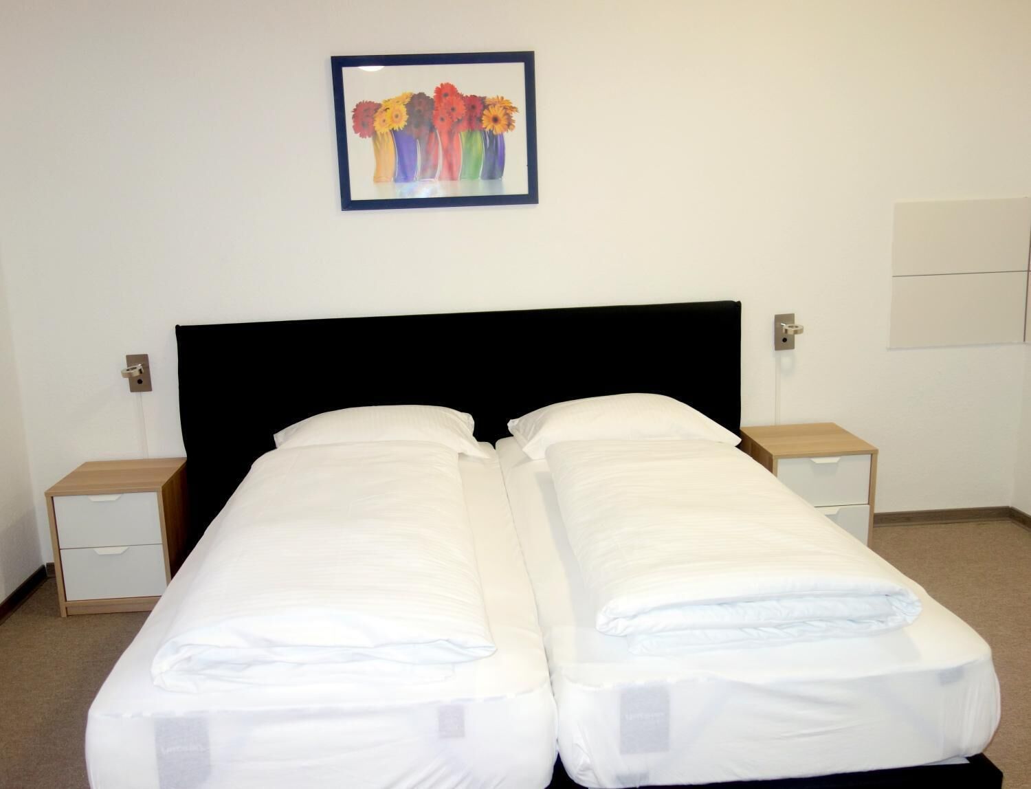 Un lit double avec du linge de lit blanc, une tête de lit noire et, au-dessus, un tableau floral encadré.