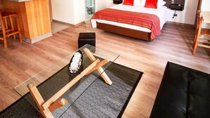 Camera d'albergo compatta con angolo cottura, un letto con biancheria rossa e bianca, un quadro appeso e un piccolo angolo salotto con tappetino e cuscini.