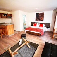 Une chambre d’hôtel compacte dotée d’une kitchenette, d’un lit avec une literie rouge et blanche, d’un tableau au mur et d’un petit coin salon avec un tapis et des coussins.