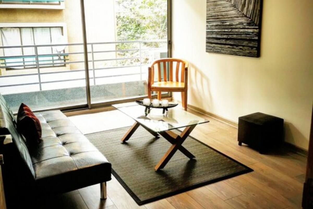 Een moderne woonkamer met een bank, een houten stoel, een glazen salontafel en een groot raam met uitzicht op groen.