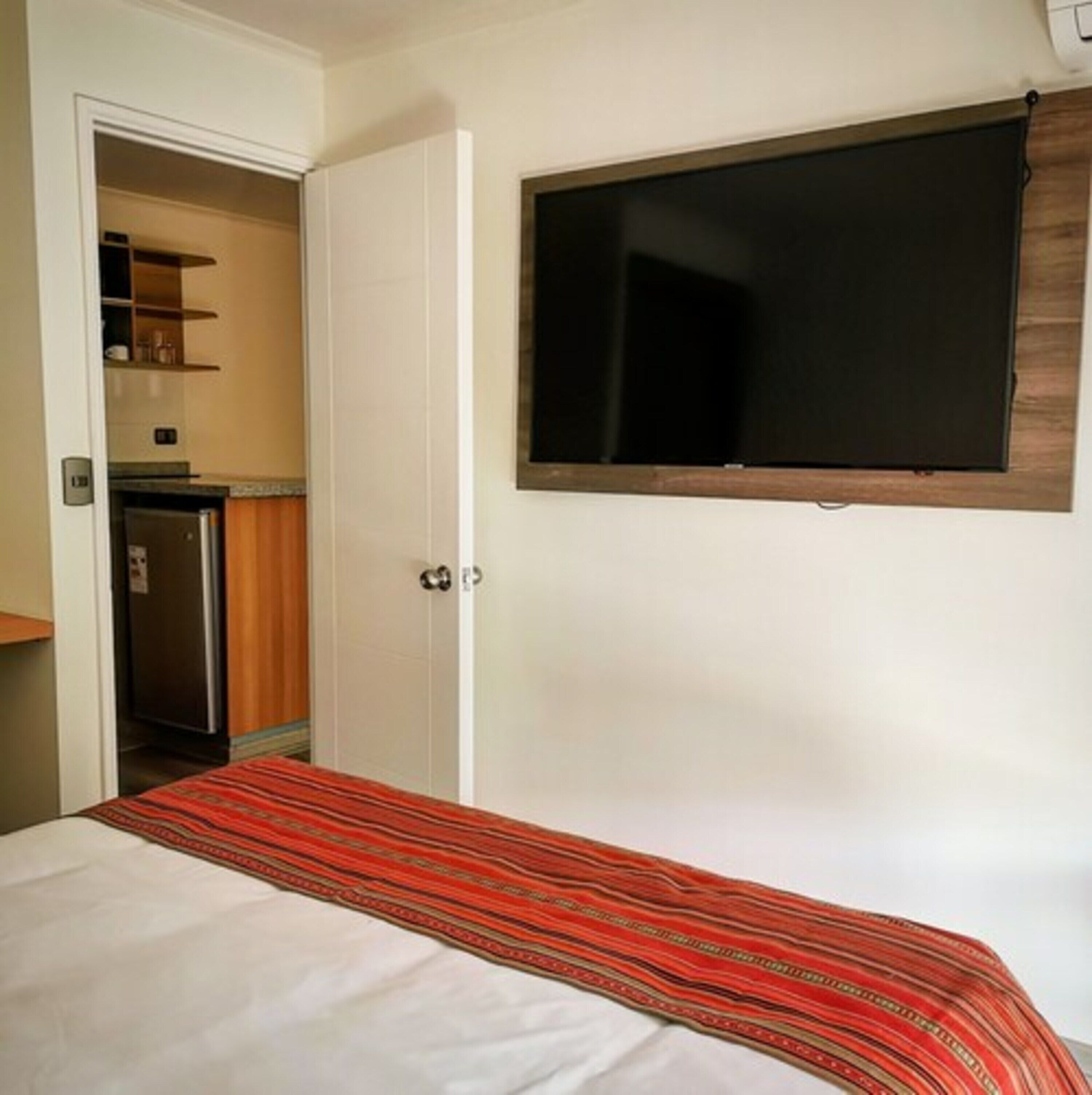 A hotel room with a bed, a television mounted on the wall, a door leading to a kitchenette, and a small shelf with items.