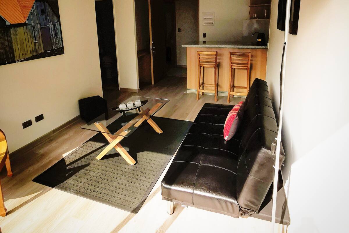 Een moderne woonkamer met een leren zwarte bank, een glazen salontafel en een schilderij aan de muur.