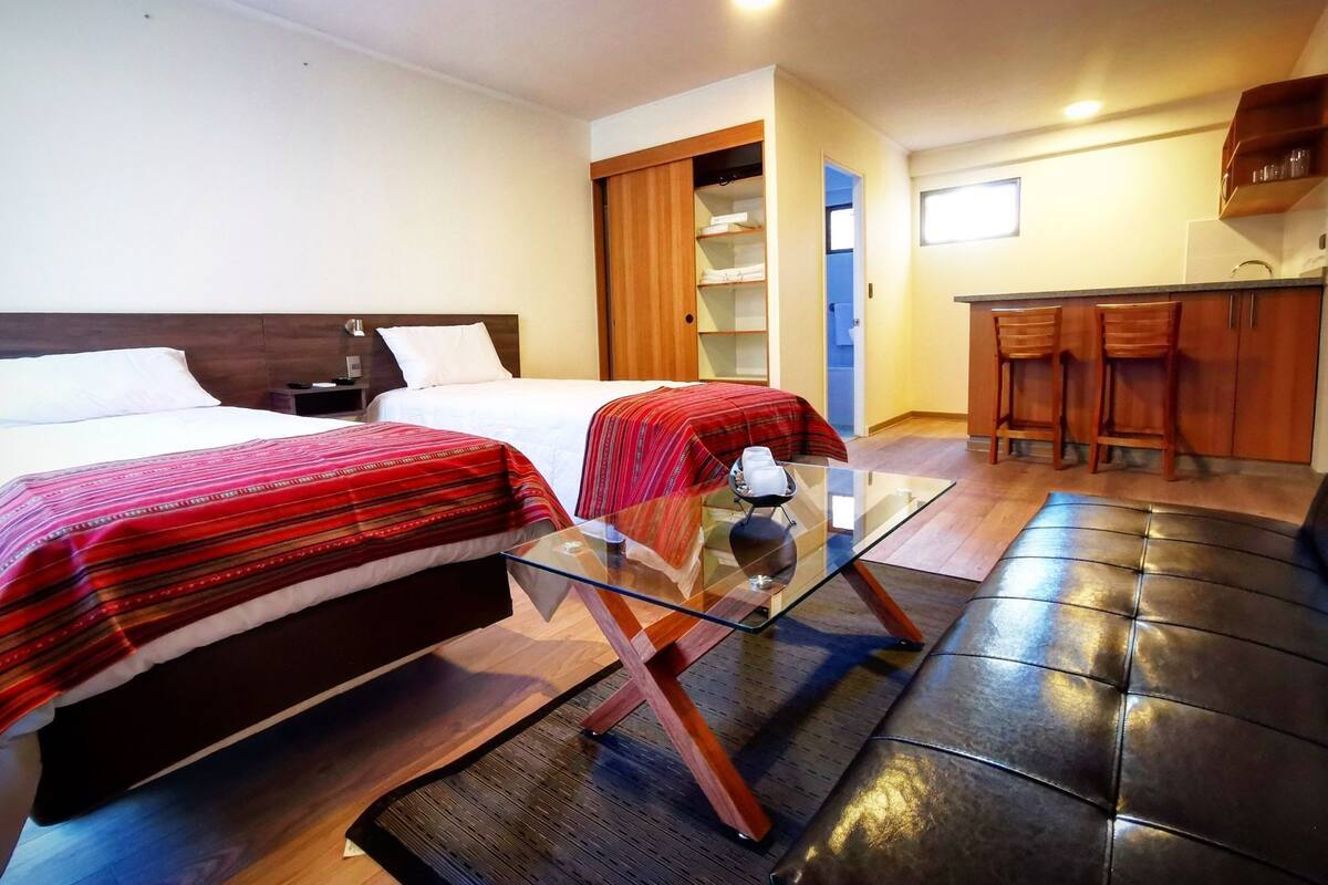 Een hotelkamer met twee bedden, een glazen tafel, een bank, een keukenhoek met barkrukken, en een badkamer die door een open deur zichtbaar is.