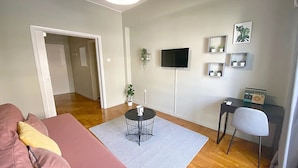 A living room with a pink sofa, a small round table with a white mug, a TV on the wall, and a radio on a side table. - Check Point - Syngrou Fix (Athens)