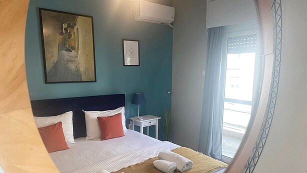 A hotel room with a bed, a nightstand, a lamp, and a window with a view. - Check Point - Syngrou Fix (Athens)