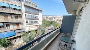 A balcony with a table and chairs, overlooking a street with buildings and trees. - Check Point - Syngrou Fix (Athens)