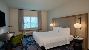 A hotel room with a bed, a desk, a chair, a window with curtains, and a nightstand with a lamp.