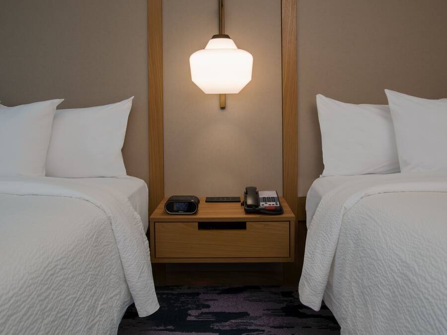 Habitación de hotel con dos camas, una mesita de noche con lámpara, teléfono y reloj digital.
