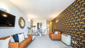 A modern living room with a sofa set, a flat-screen TV, a decorative mirror, and a patterned wallpaper. - Modern 2 bedroom in Kensington (London)
