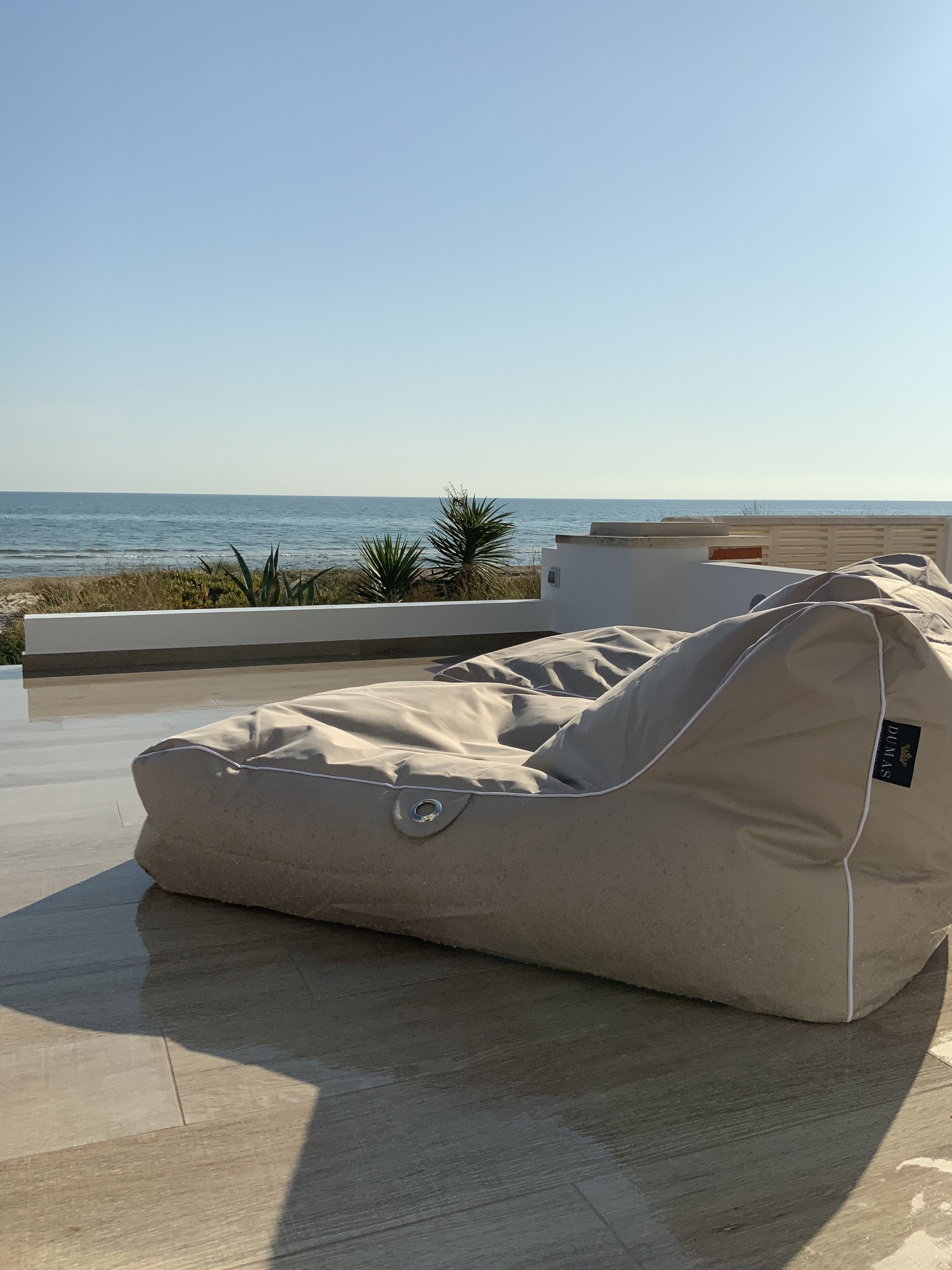 Un área de estar exterior en tonos beige, con un aro metálico de manija, con vista a una playa y al océano, con vegetación de fondo.