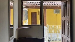A narrow hallway with a bed, a ceiling fan, a television mounted on the wall, and a door leading to an outdoor area with a yellow wall and a white railing. - Pousada Florescer (Paraty)