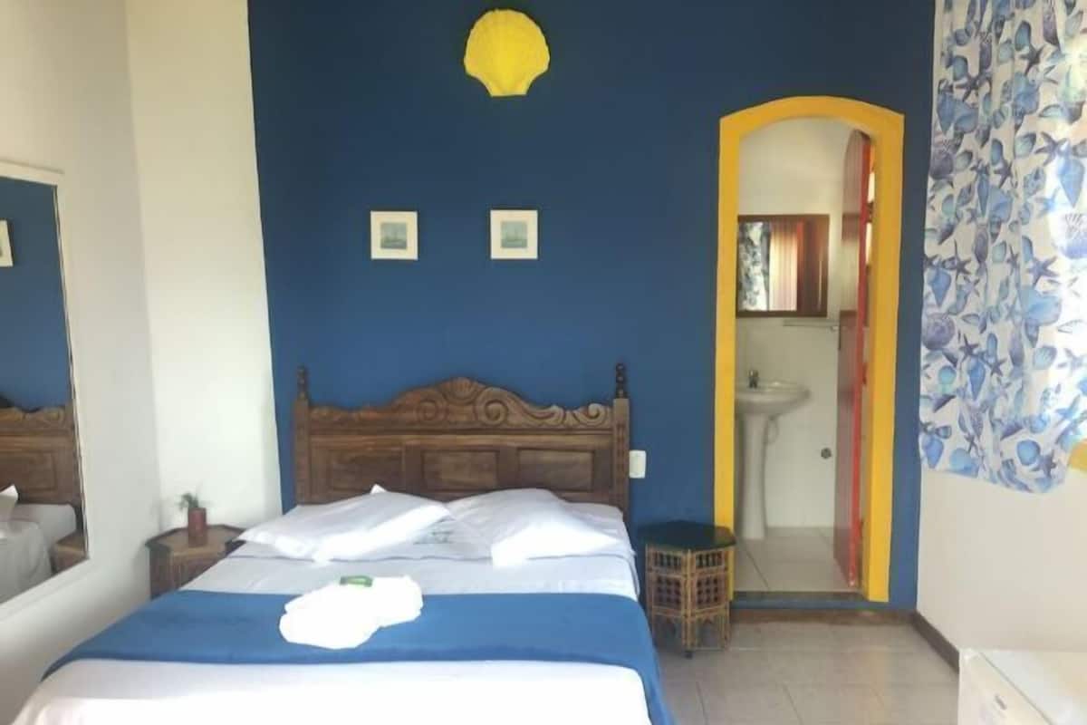A bedroom with a wooden bed, a small bedside table, a mirror, and a bathroom visible through an archway.
