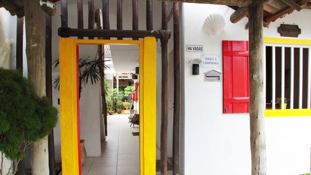 A building entrance with a yellow and red door, a sign reading "HA VAGAS", and a window with a red shutter. - Pousada Florescer (Paraty)