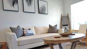 A living room with a white sofa, patterned cushions, a round wooden coffee table, and three framed leaf artworks on the wall.