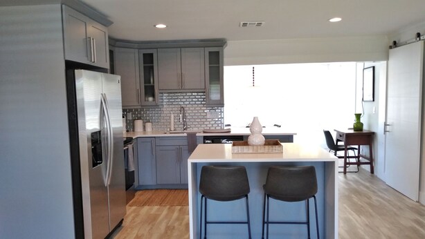 A modern kitchen with grey cabinets, a stainless steel refrigerator, a wooden island with two bar stools, and a dining area with a table and chairs.