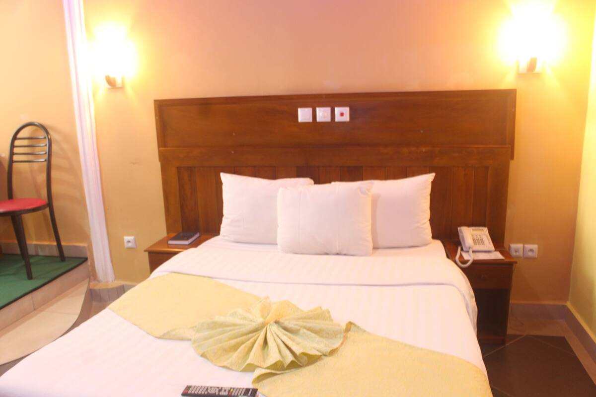 A hotel room with a wooden headboard, a bed with white linens, a bedside table with a telephone, and a chair with a red cushion.