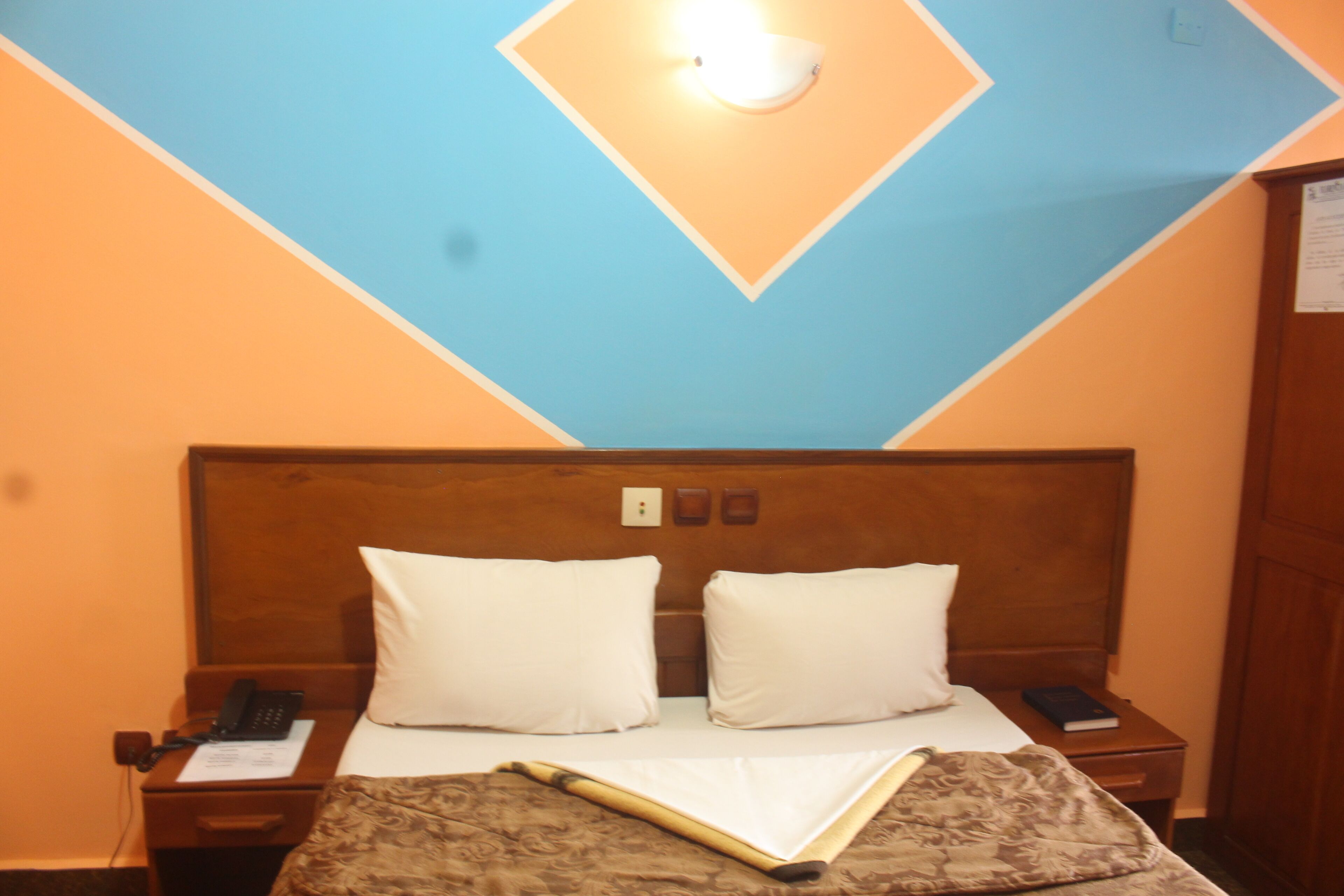 A hotel room with a wooden headboard, two white pillows, a bedside table with a phone and a book, and a patterned bedspread.