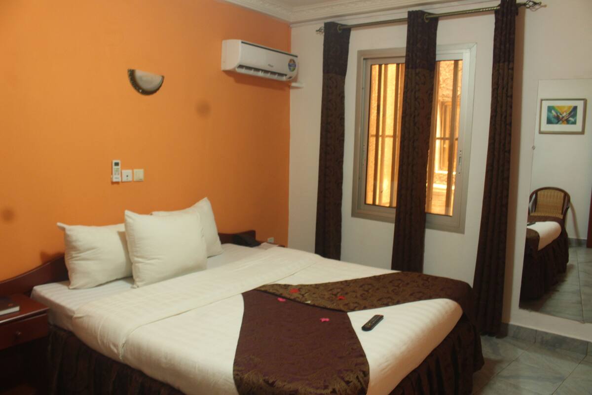 A hotel room with a bed, an air conditioning unit, a window with curtains, and a chair.
