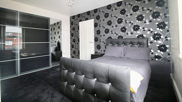 A bedroom with a tufted headboard, a bed with grey bedding, a wardrobe with mirrored doors, and a floral-patterned wall.