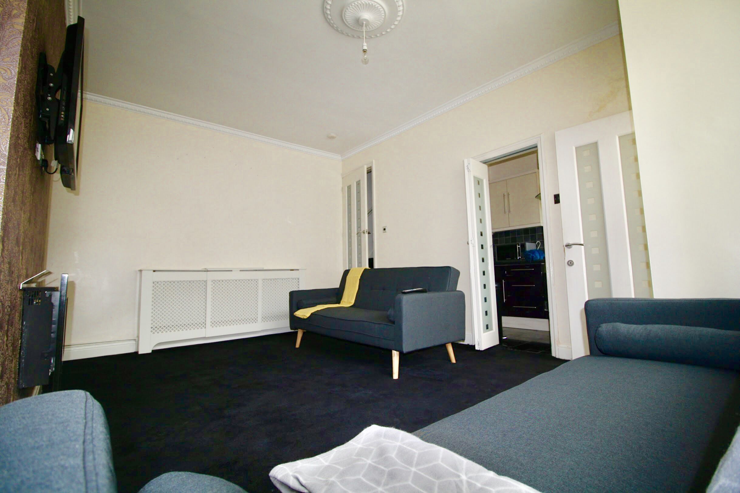 Una sala moderna con un sofá gris, un sofá negro y un sillón gris. Hay un televisor montado en la pared y un radiador blanco debajo de la ventana.