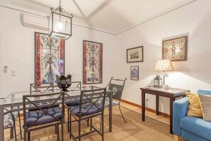 Uma área de refeições com mesa de vidro e cadeiras, um sofá azul e obras de arte emolduradas nas paredes.