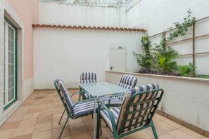 Um pequeno pátio com uma mesa e cadeiras, uma parede branca e piso ladrilhado.