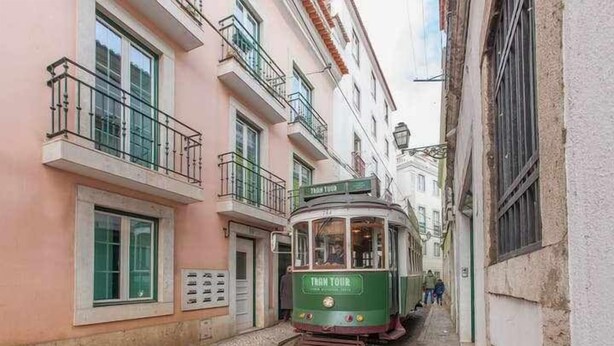 A green tram with "TRAM TOUR" written on it, traveling down a narrow street flanked by buildings with balconies and windows. - Elegant in Alfama with Terrace and FREE parking. (Lisboa)
