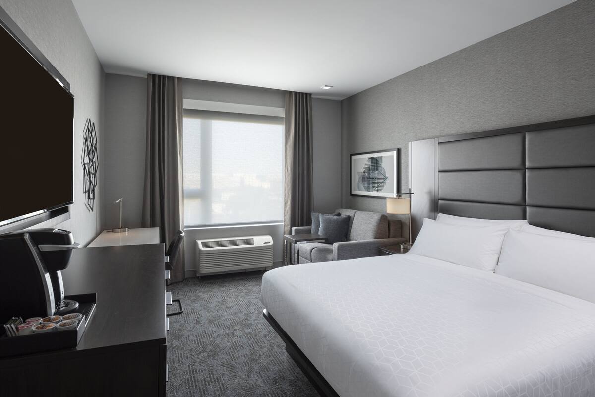 A modern hotel room with a large bed, a desk with a coffee machine, a chair, a TV, and a large window with curtains.