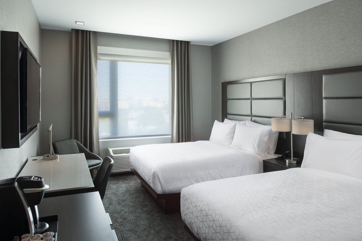 Quarto de hotel com duas camas, uma escrivaninha, uma cadeira, uma televisão e uma janela grande com cortinas.