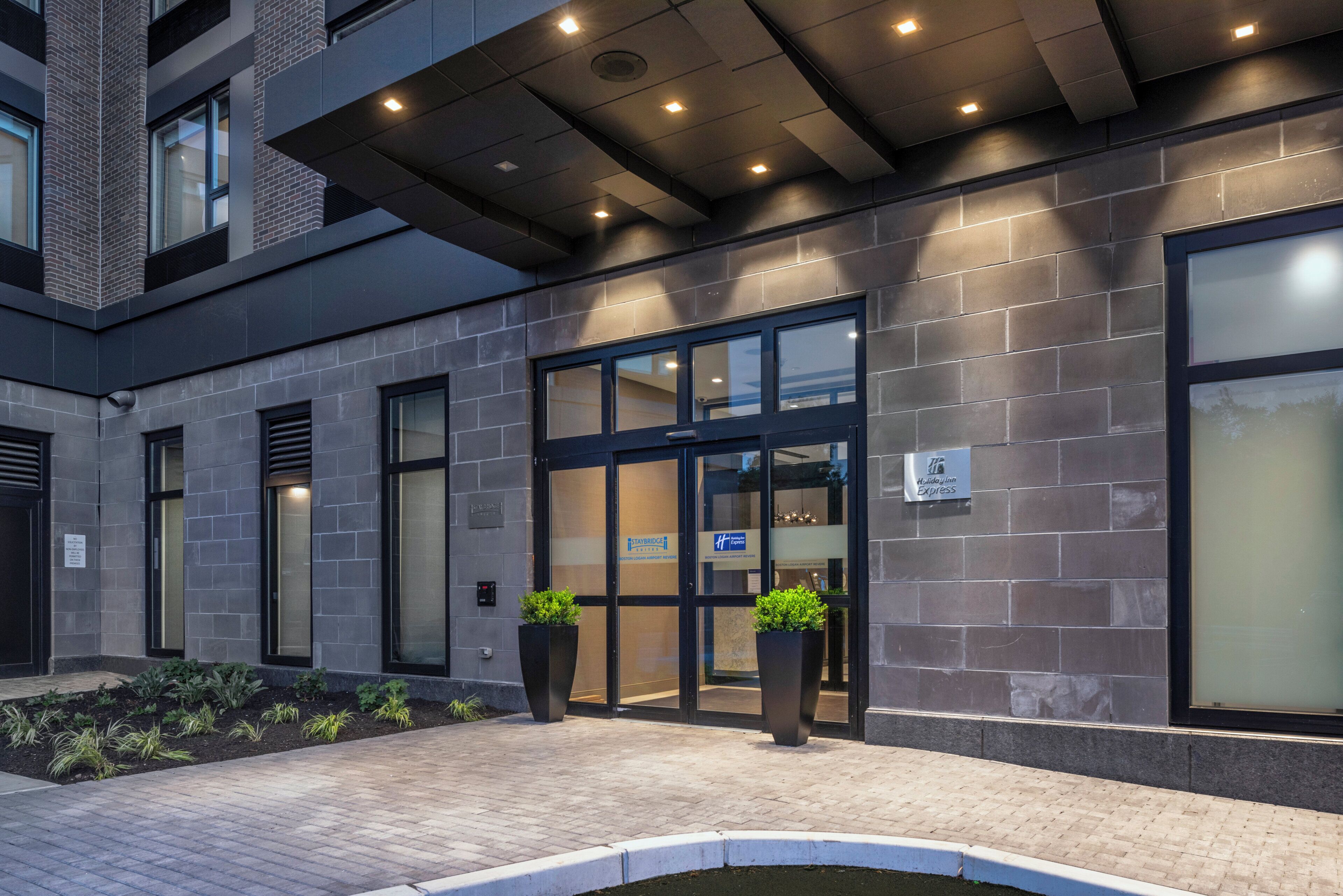 A hotel entrance with a modern design, featuring a brick facade, large windows, and potted plants.