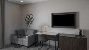 A modern hotel room with a grey sofa, a desk with a chair, a coffee machine, and a TV mounted on the wall. - Holiday Inn Express Boston Logan Airport - Revere by IHG (Revere)