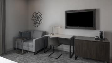 A modern hotel room with a grey sofa, a desk with a chair, a coffee machine, and a TV mounted on the wall.