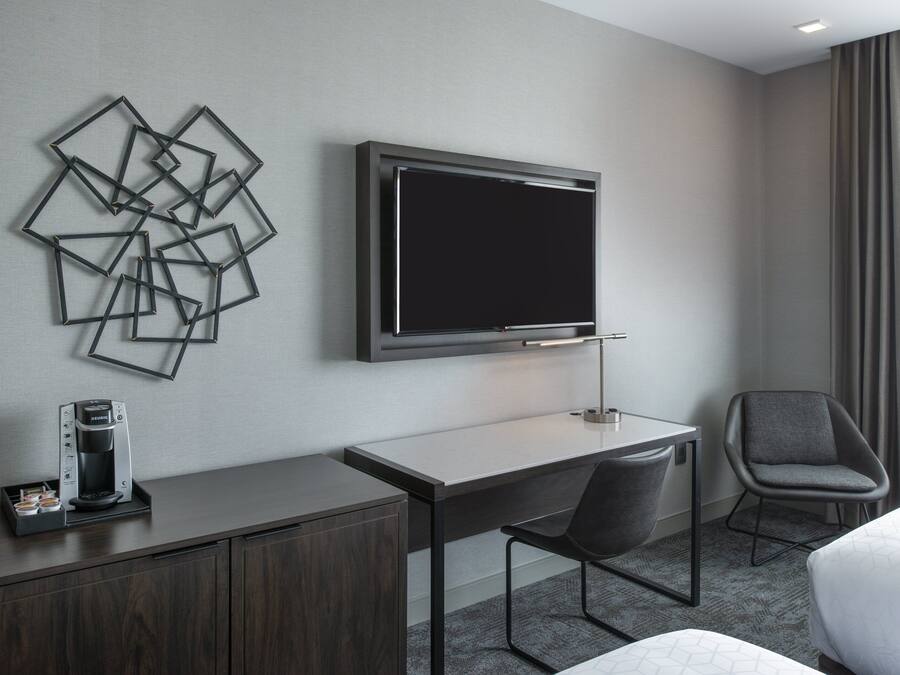 Quarto de hotel com televisão de tela plana na parede, uma escrivaninha com cadeira, cafeteira e cama com edredom branco.