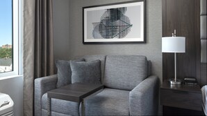 A modern hotel room with a grey sofa, a small table, a framed abstract artwork, a bedside lamp, and a view of the city through the window. - Holiday Inn Express Boston Logan Airport - Revere by IHG (Revere)